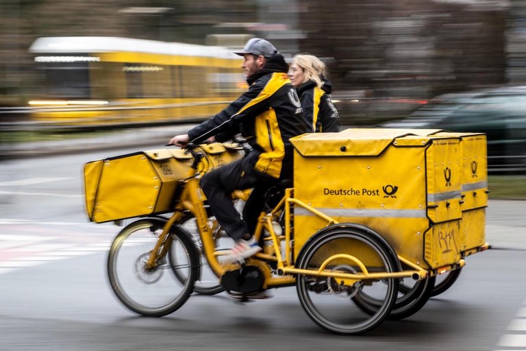 Eine Briefzustellerin und Briefzusteller auf ihren Zustellfahrrädern im Regen