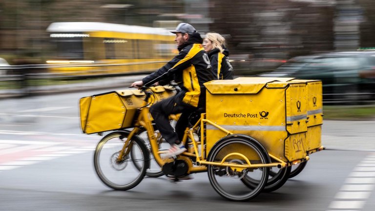 Eine Briefzustellerin und Briefzusteller auf ihren Zustellfahrrädern im Regen