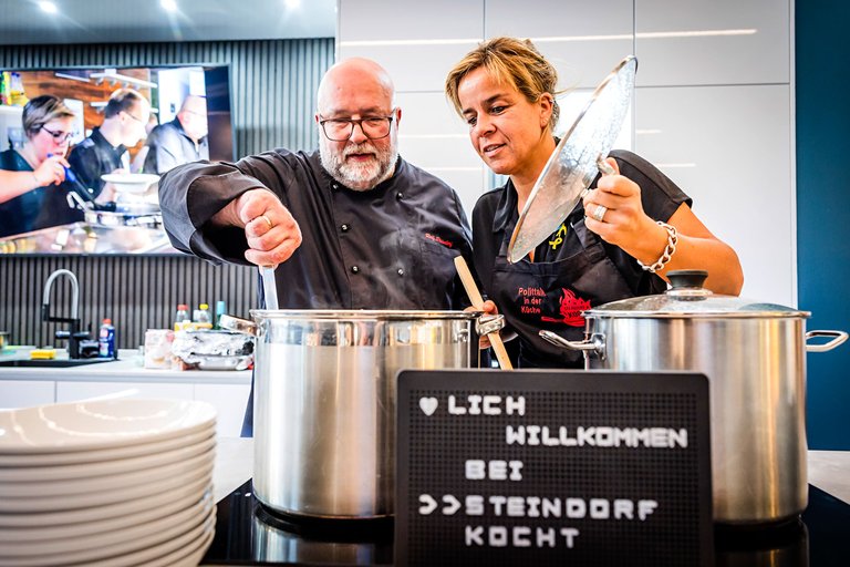 Steindorf und Neubaur stehen gemeinsam am Herd und schauen in die großen Kochtöpfe