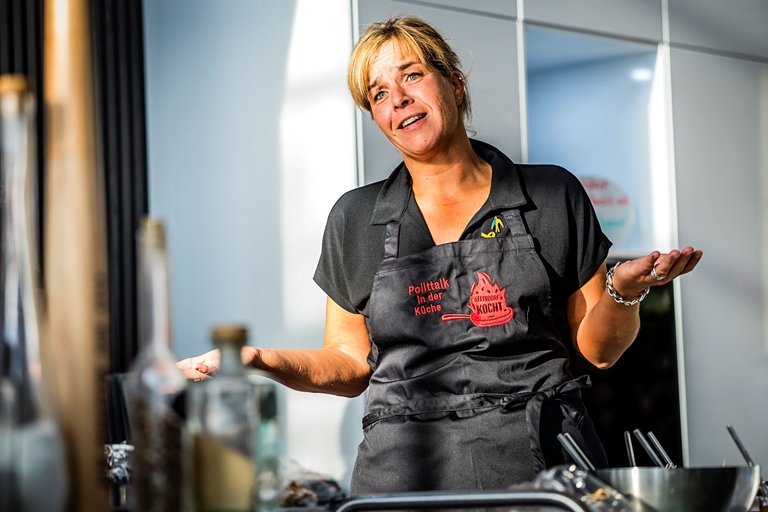 NRW-Wirtschaftsministerin und stellvertretende Ministerpräsidentin Mona Neubaur redet und gestikuliert. Sie trägt eine Schürze mit der Aufschrift "Politiktalk in der Küche"